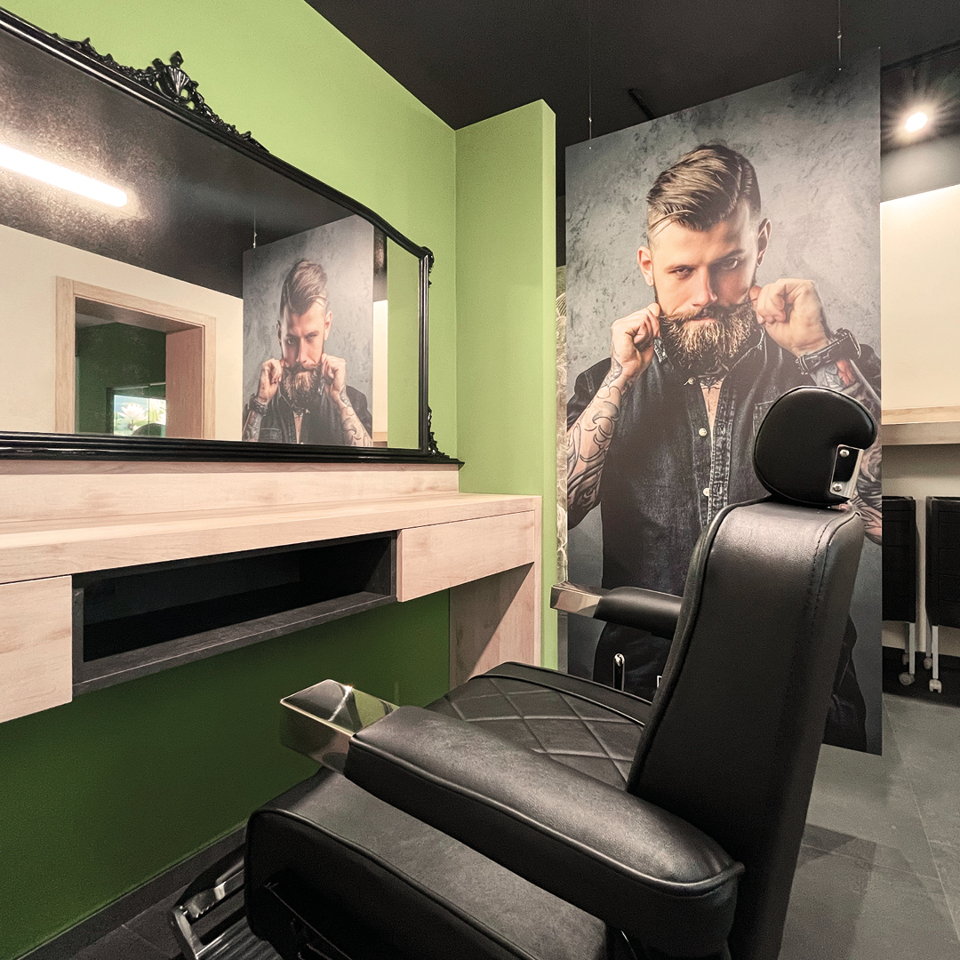 Immagine di una postazione dell'area barber con poltrona  nera, gramde specchio rettangolare e pareti verdi decorate con immagine di uomo 
